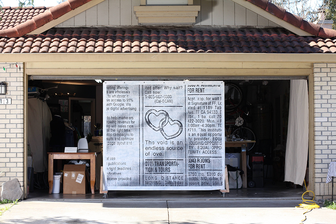 A suburban garage with an open garage door, framing a large wall with a large print of a scanned classified ad featuring two interlocked hearts and text reading the void is an endless source of love.