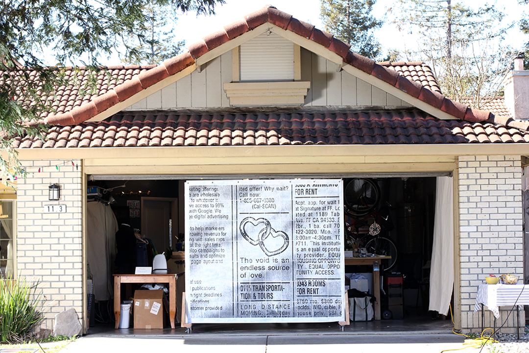 A suburban garage with an open garage door, framing a large wall with a large print of a scanned classified ad featureing two interlocked hearts and text reading the void is an endless source of love.