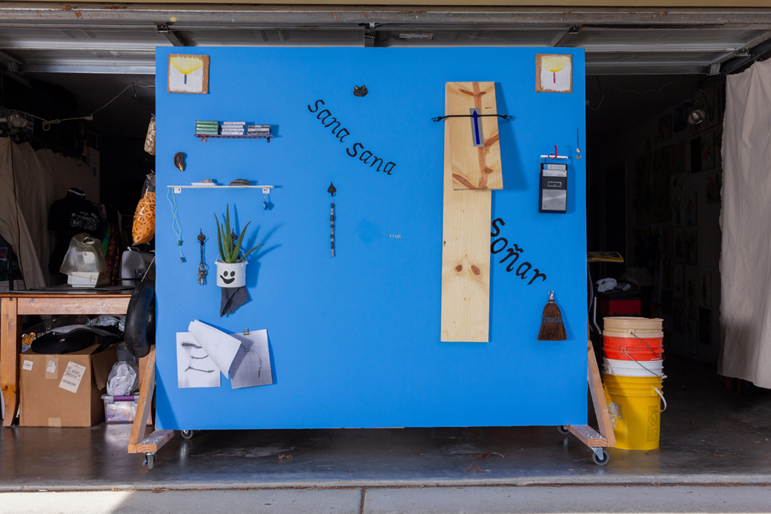 A blue wall with several objects including a wooden bench, silver milagros, a cassette player, paintings of candles, an aloe plant, tools, and more.