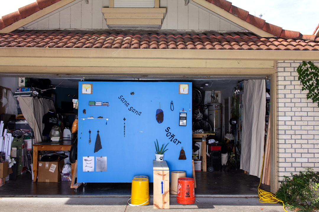 A blue wall with several objects including a wooden bench, silver milagros, 
a cassette player, paintings of candles, an aloe plant, tools, and more..