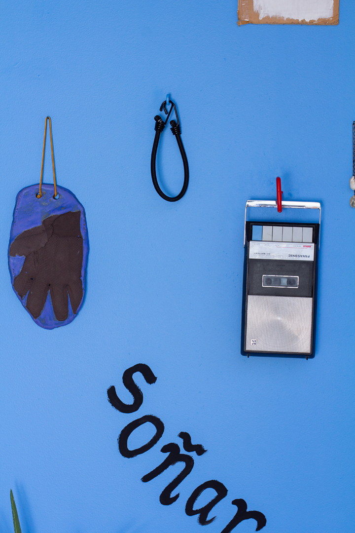 A blue wall with several objects including a wooden bench, silver milagros, a cassette player, paintings of candles, an aloe plant, tools, and more.