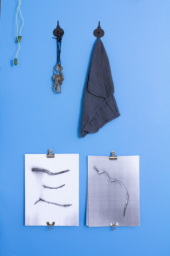 A blue wall with several objects including a wooden bench, silver milagros, a cassette player, paintings of candles, an aloe plant, tools, and more.