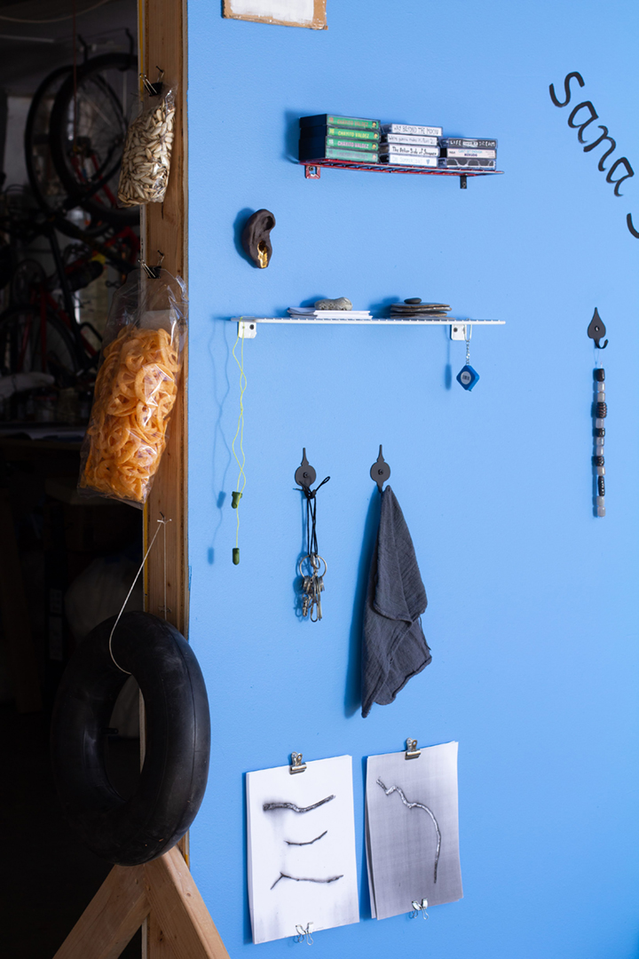 A blue wall with several objects including a wooden bench, silver milagros, a cassette player, paintings of candles, an aloe plant, tools, and more.