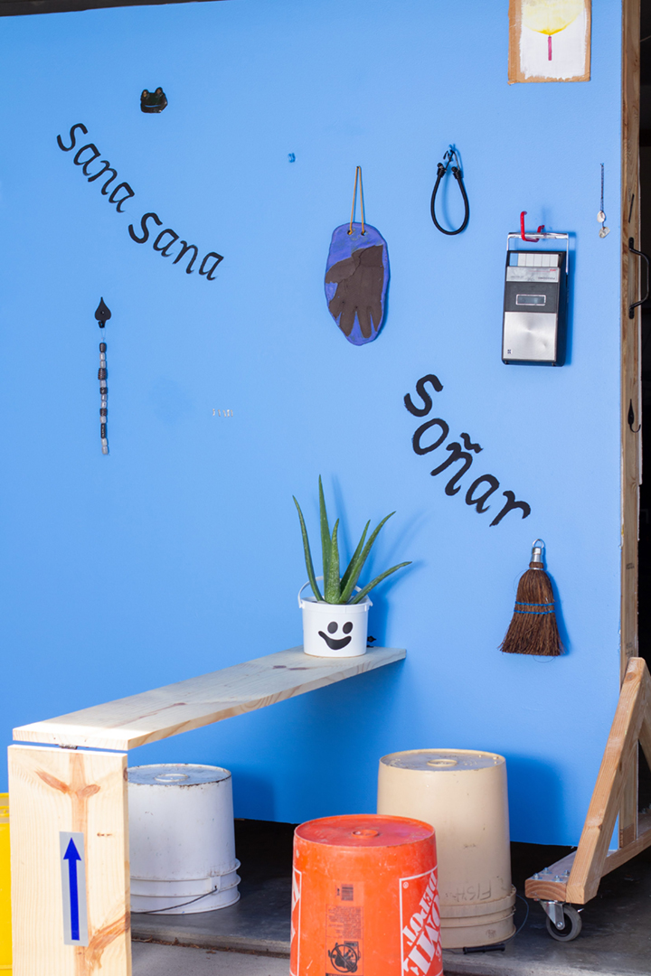 A blue wall with several objects including a wooden bench, silver milagros, a cassette player, paintings of candles, an aloe plant, tools, and more.