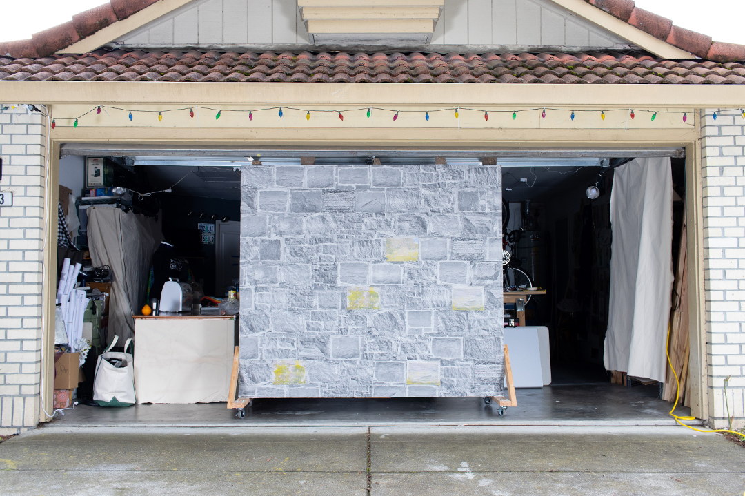 A suburban garage with an open garage door, framing a large wall papered with wheatpasted images of stone from stone walls.