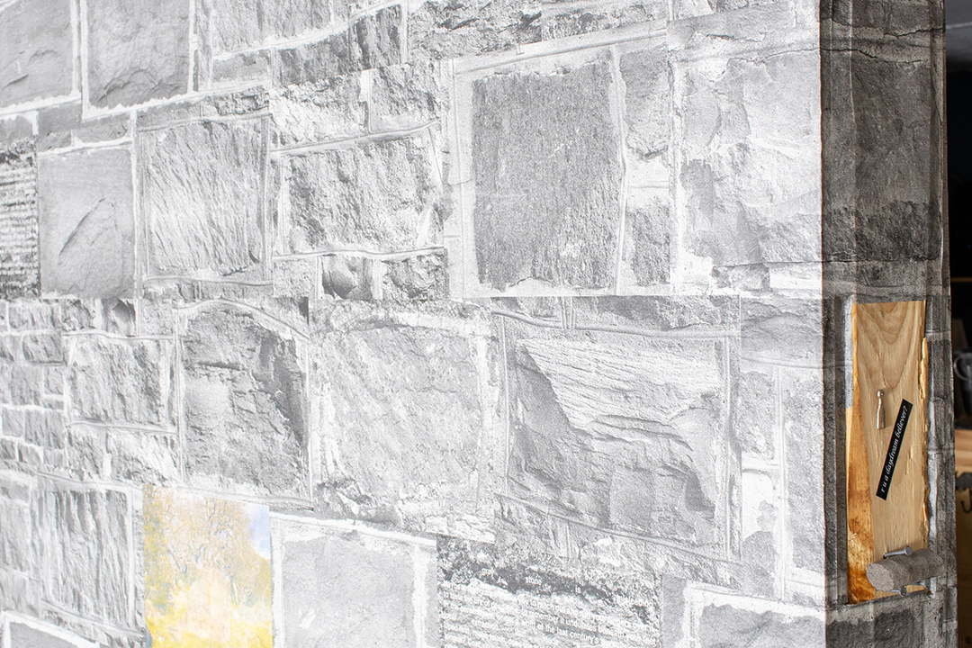 A view of the side edge of the large wall papered with wheatpasted images of stone from stone walls, where a small piece of stone is attached to the bare wood of the wall, along with a small metal charm and a piece of black label