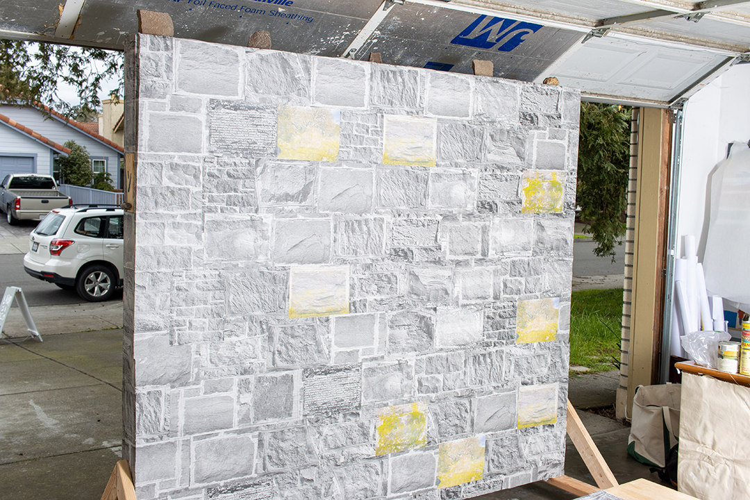 The backside of a large wall papered with wheatpasted images of stone from stone walls in an open garage door, the street visible beyond.