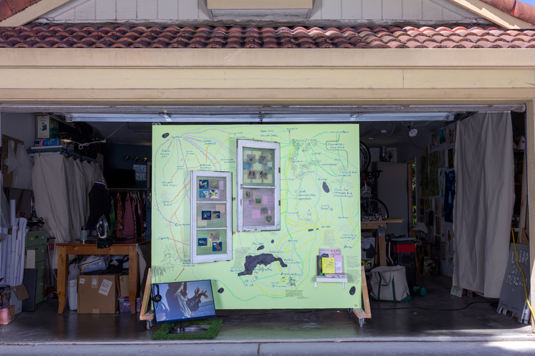 A suburban garage with an open garage door, framing a large wall with a painted diagram of words and images and hung with two window frames. In front of it is a small television monitor with an image of people.